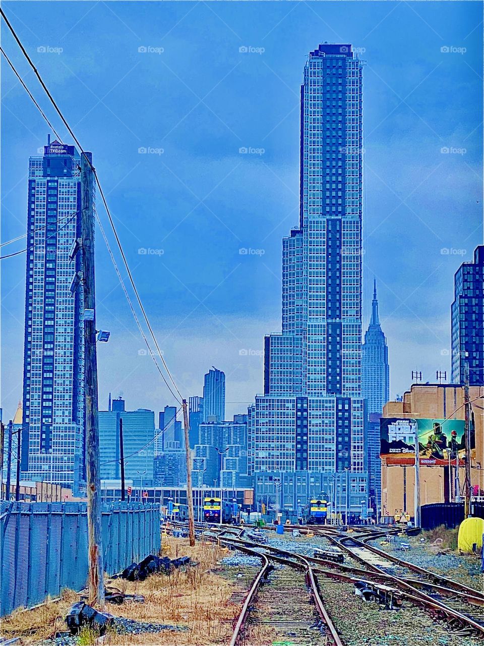 Urban high rises seen at the far end of the train tracks on Municipal Transit Authority grounds pedestrians have to cross in order to ascend the red metal staircase leading up onto the Pulaski Bridge in LIC, Queens. 2021. Hypnotic Productions