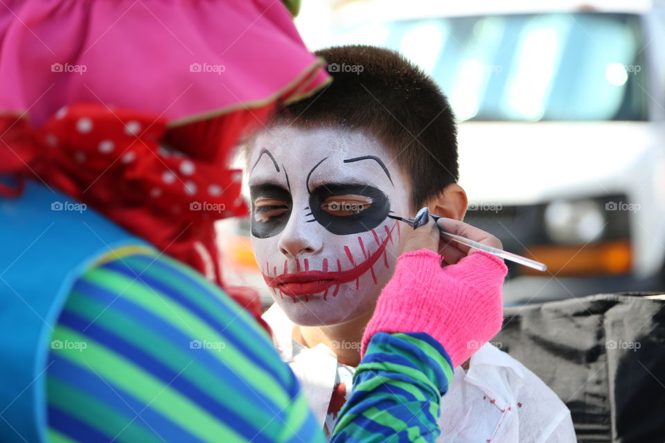 Colorful clown applying face paint designs to young boy. Mask is white base, red stitched mouth, black eyes