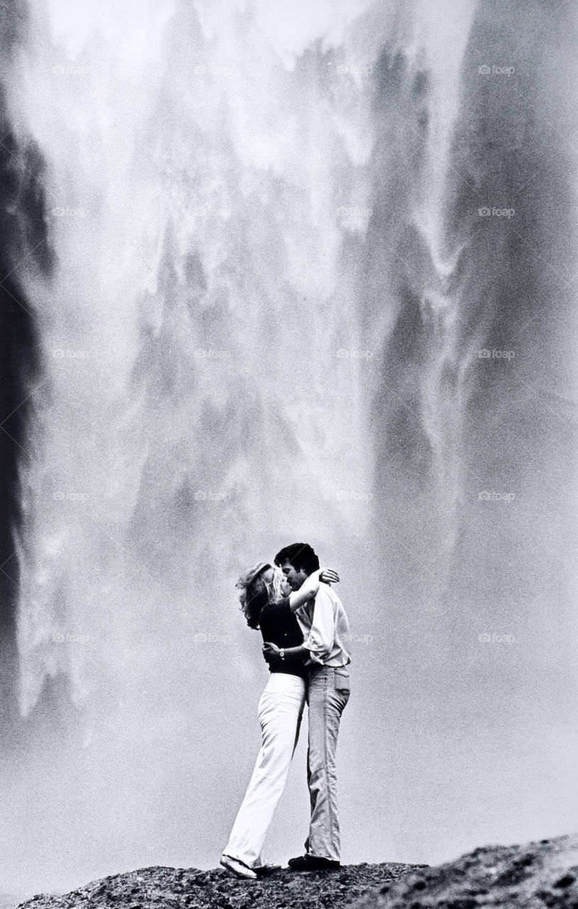 Young lovers kiss in front of an alpine waterfall.