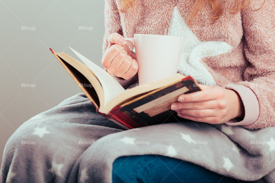 Girl enjoying the reading a book and drinking coffee at home. Young woman sitting on a chair, wrapped in blanket, holding book, relaxing at home. Portrait orientation. View from above