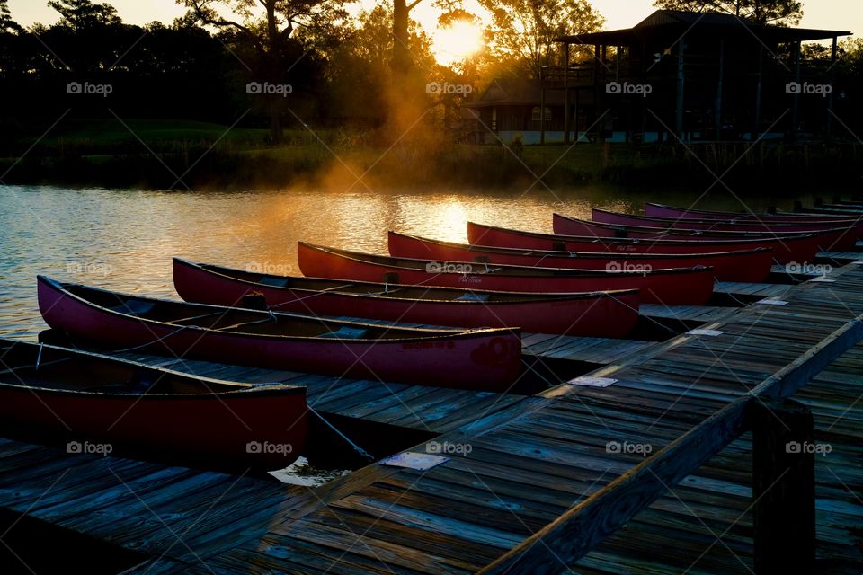 Beautiful row of docked red canoes on a lake in the early morning sun at a park in Arapahoe North Carolina. Gentle fog moving over the water backlit by the rising sun. 