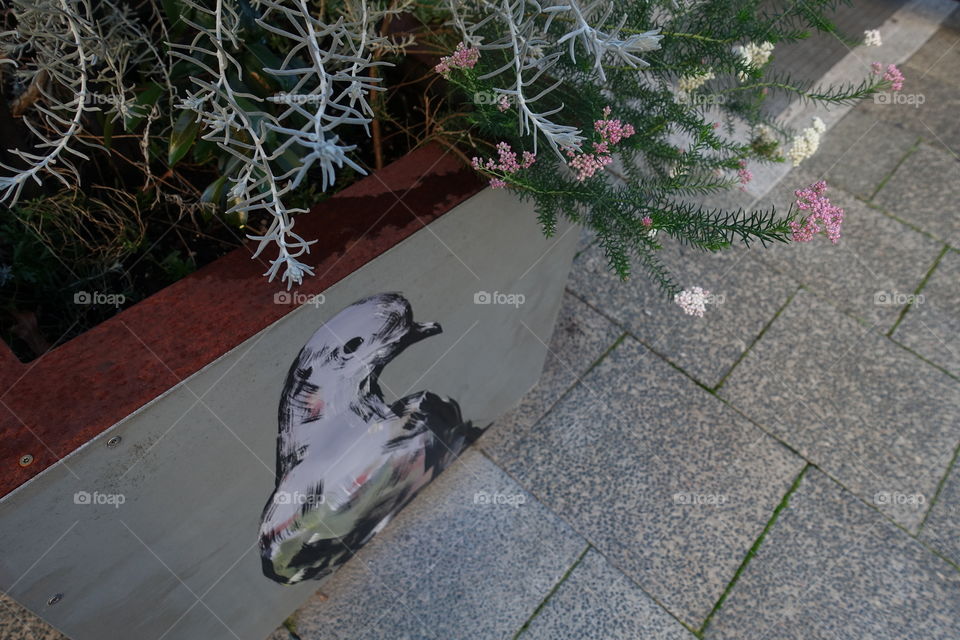 Small flowers are in the plant pot painted pigeon artwork.  The artwork is situated on the street.