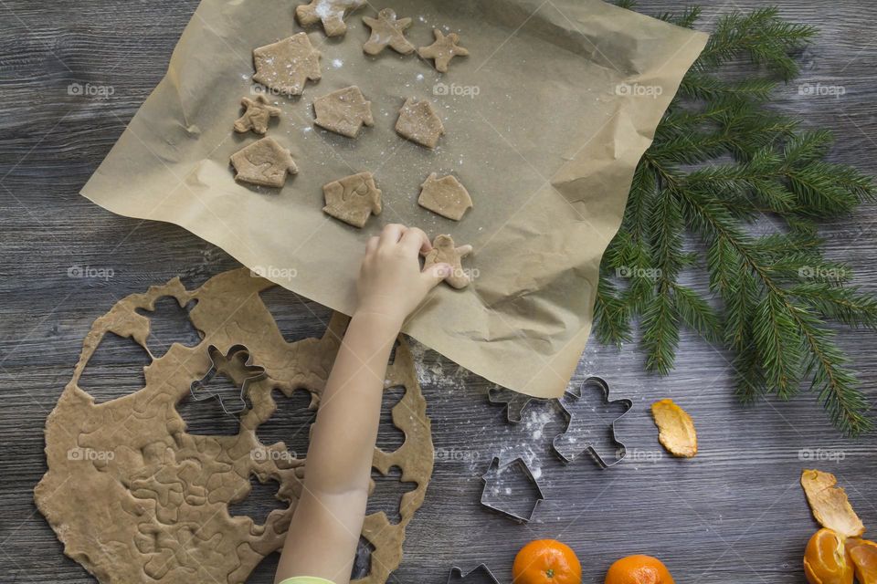 Little child helps cooking christmas ginger cookies on a wooden table with tangerines and green Christmas trees.