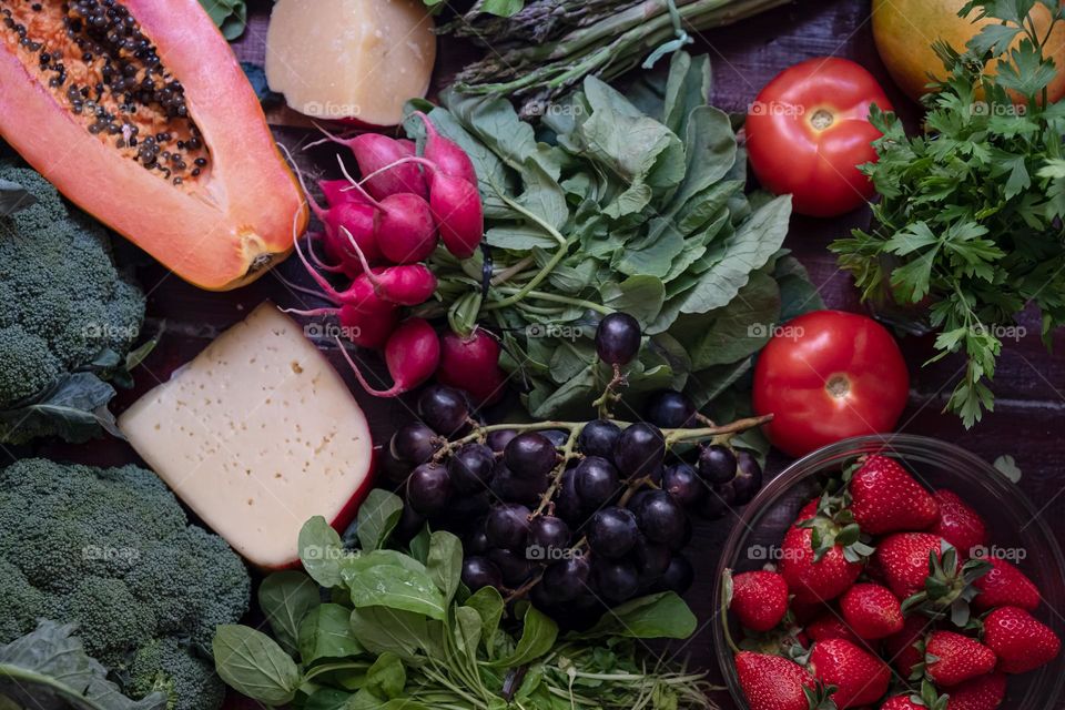 A wooden table full of vegetables, herbs, berries, fruits and cheese. Flat lay with cut papaya, strawberry, broсcoli, arugula, tomatoes, mango, cilantro, radish, cheese, Parmesan