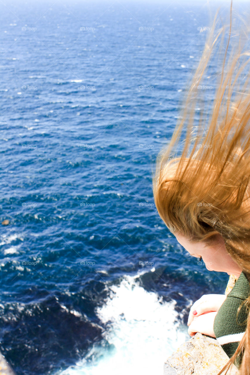Girl looking off of a cliff into the ocean as the wind blows back her hair