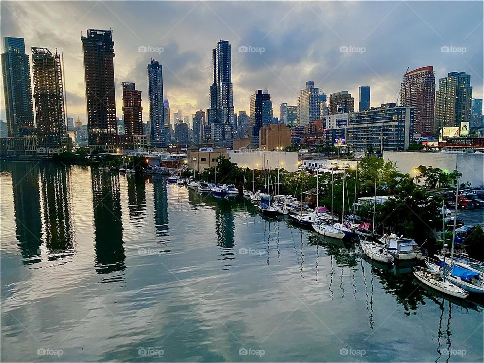 This is “Newtown Creek” with its many different kinds of boats seen from the “Pulaski Bridge” in LIC, Queens at twilight time just before nightfall. The sun’s last setting rays can be seen reflected in the “East River”. 2023. Hypnotic Productions