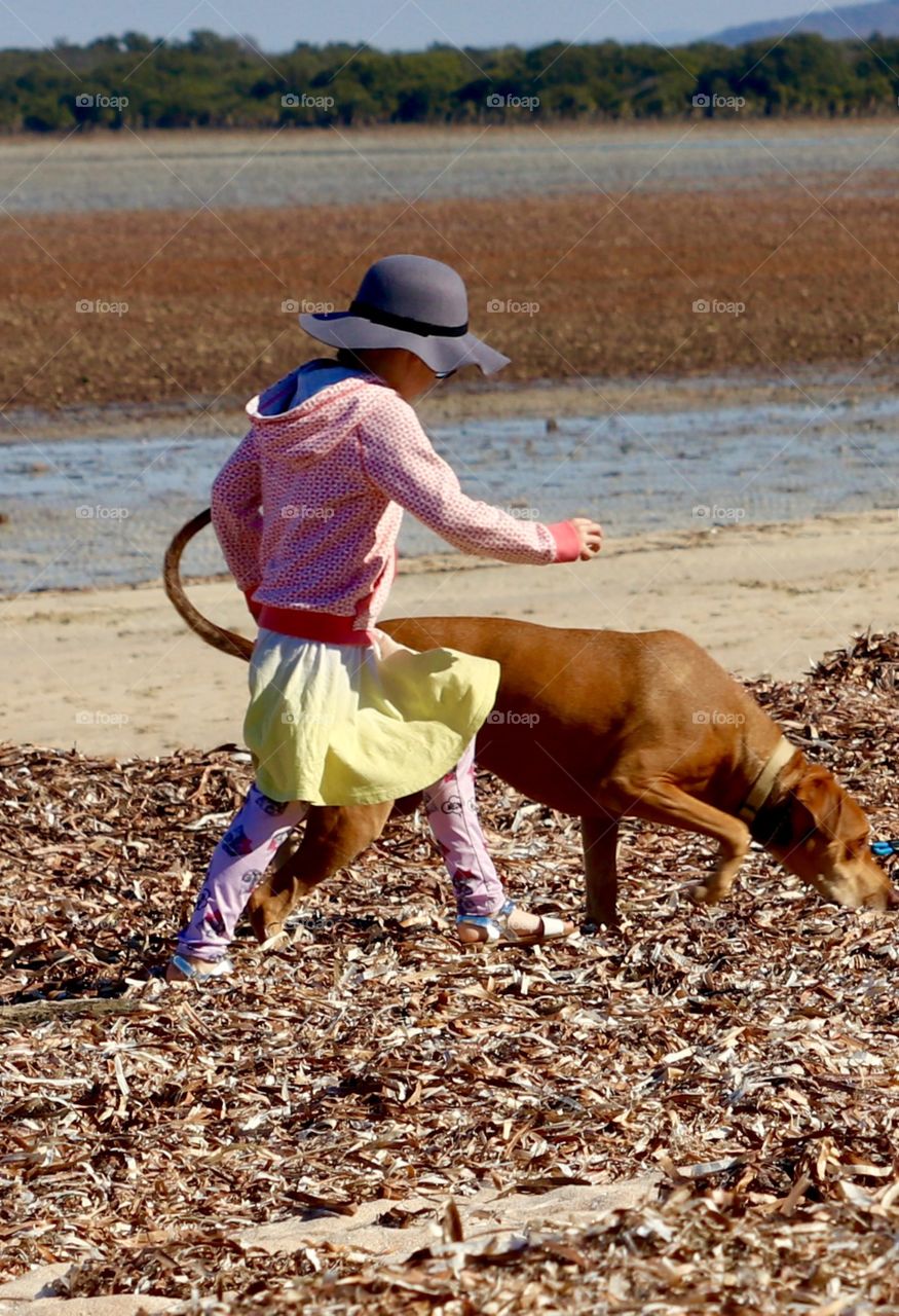 Little girl in yellow skirt, pink jacket, hat and leggings running on beach with pet dog