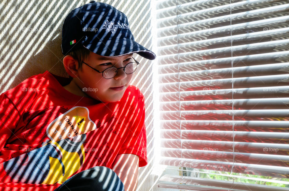 A boy with eyeglasses sits by the window. The sun shines through the blinds.