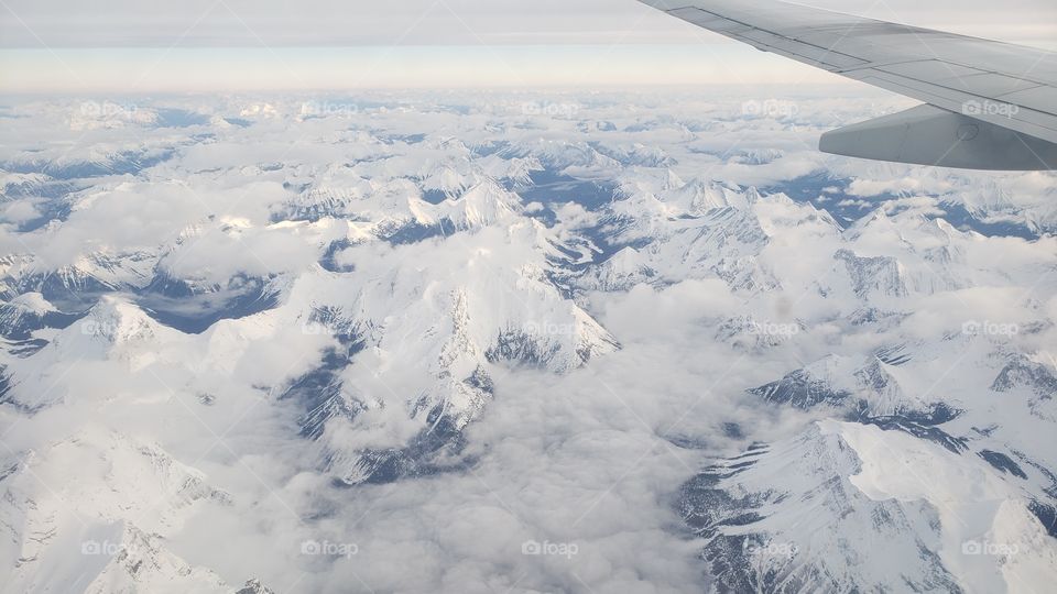 View from above, looking out of airplane window while flying over snow covered Canadian rockies during cloudy winter day.