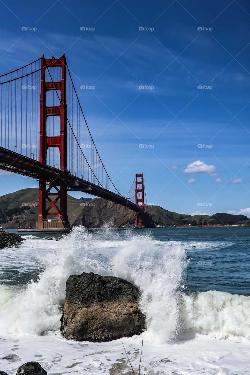Golden Gate Bridge viewed from Fort Point in San Francisco California with the waves crashing onto the rocks and the clear blue skies above