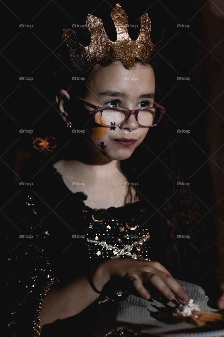 Portrait of a beautiful caucasian girl in a golden crown, glasses and a black dress of a dead princess eating pooworn with taoelkii looking attentively to the side at night at a school holiday on the street, side view close-up.
Halloween celebration