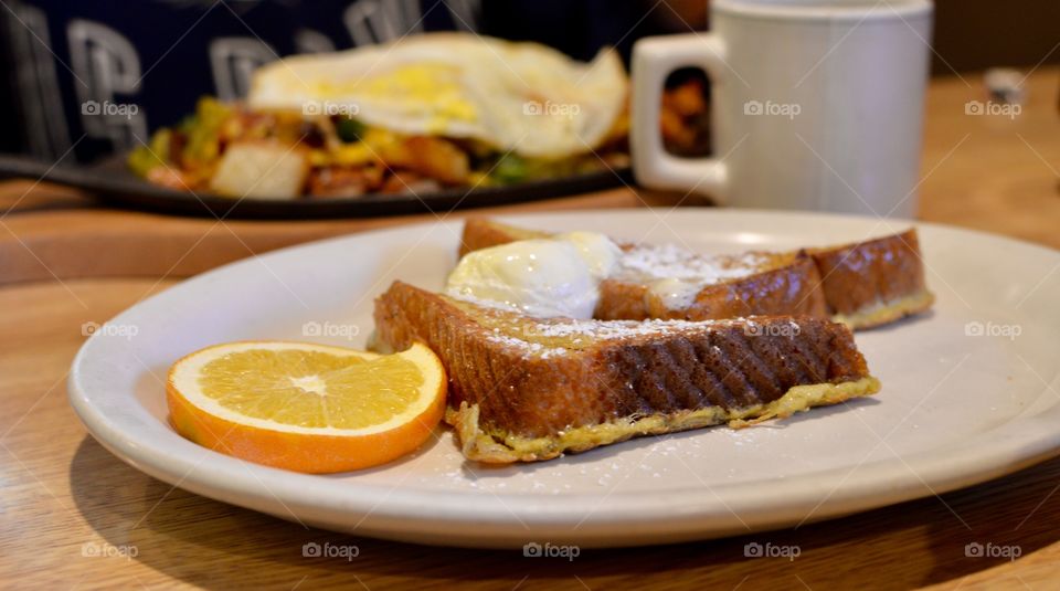 Two delicious slices of French toast on a plat with an orange slice topped with butter and powdered sugar 