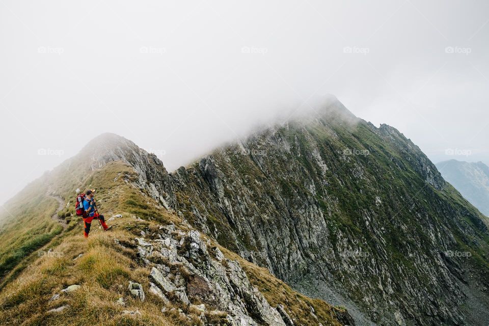 Man standing at the edge of a mountain ridge, while hiking on a cloudy day at the end of the summer.