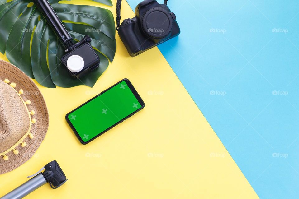 Photographer's set of camera, action camera, selfie stick, green screen smartphone, straw hat and manster leaf lie on the left on a blue-yellow background with copy space space, flat lay close-up.