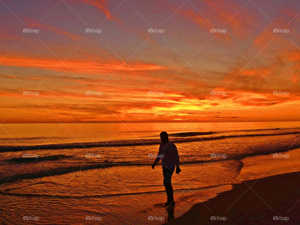 
A man walking along the beach in front of the Gulf of Mexico. The color of twilight and sunset. The dramatic hurried rush of orange collapse as the sun sinks below the gorgeous, cloudy skyline