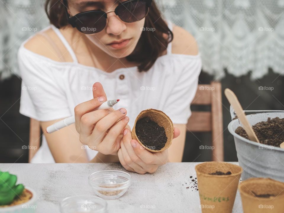 Portrait of a beautiful caucasian brunette teenage girl with sad emotion and a disgruntled face sits at a light stone table and writes in flamaskrom on a cardboard glass with earth and seeds, close-up side view. The concept of planting seeds,spring w