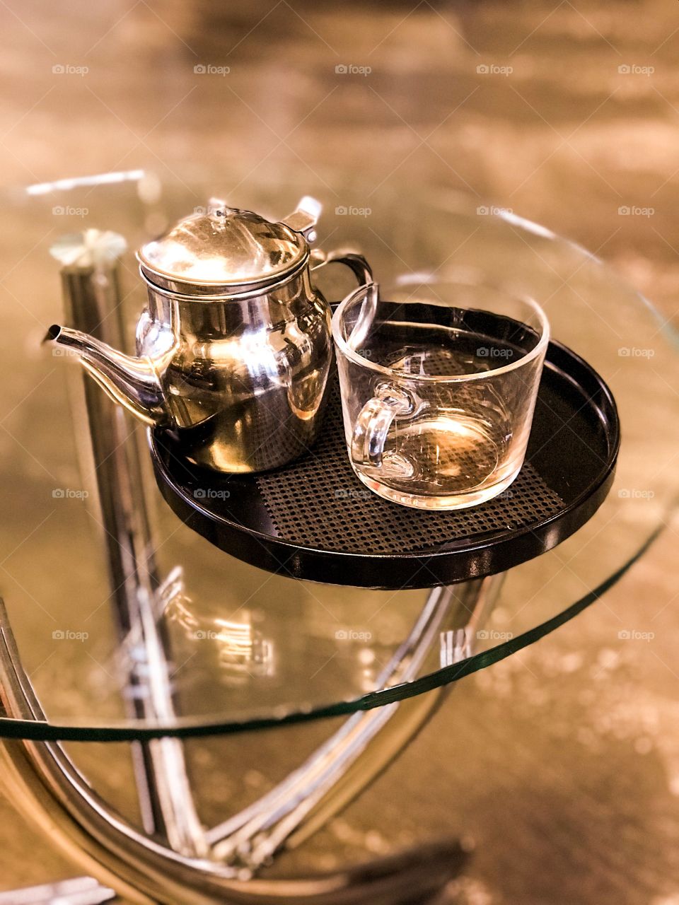 The light is reflected off multiple surfaces at multiple angles; glass teacup, glass table, stainless steel teapot, chrome table legs. The warm light from the filtered spotlights produces a warm glow complemented further by the brown stone floor.