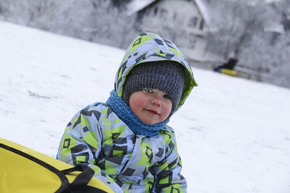 A small, carefree boy walks in the white snow in winter and rides a tubing in the park, near trees in the snow.
