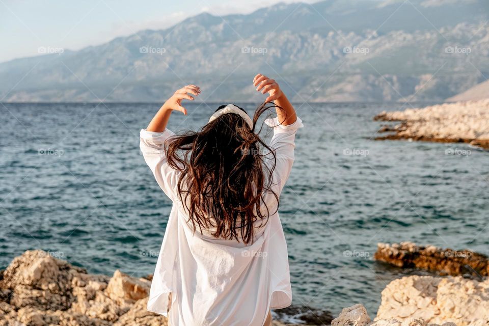 Back view of a young woman in a white shirt standing on the rocky coast of the sea