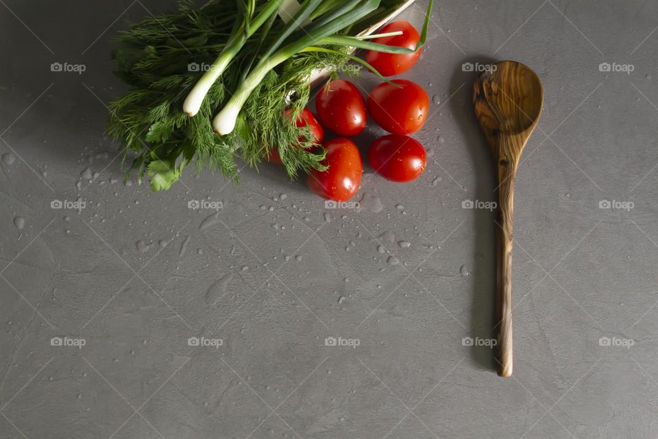 Fresh, bright, juicy vegetables, a bamboo spoon and greens for salads and other vegetable dishes are on a gray kitchen worktop.