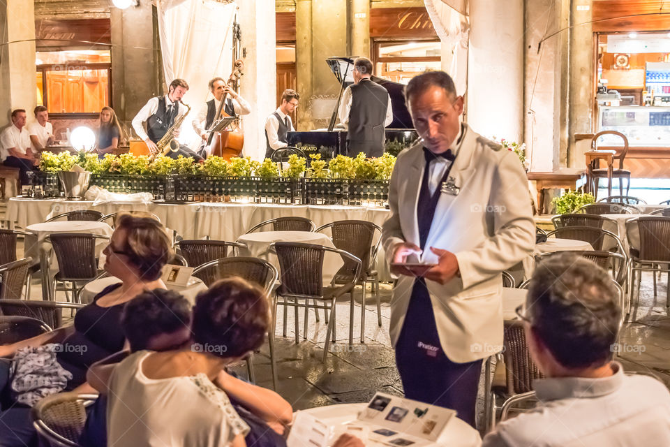 Waiter With Tablet Taking Order From Customers In An Outdoor Restaurant With A Live Music Jazz Band In Venice, Italy
