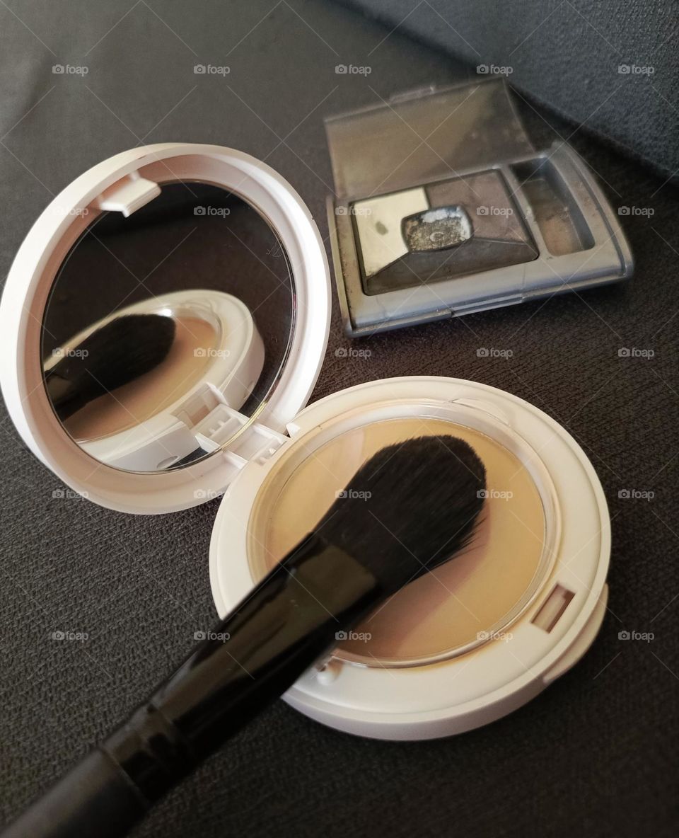 Compact powder, powder brush and eyeshadow with dark grey background.