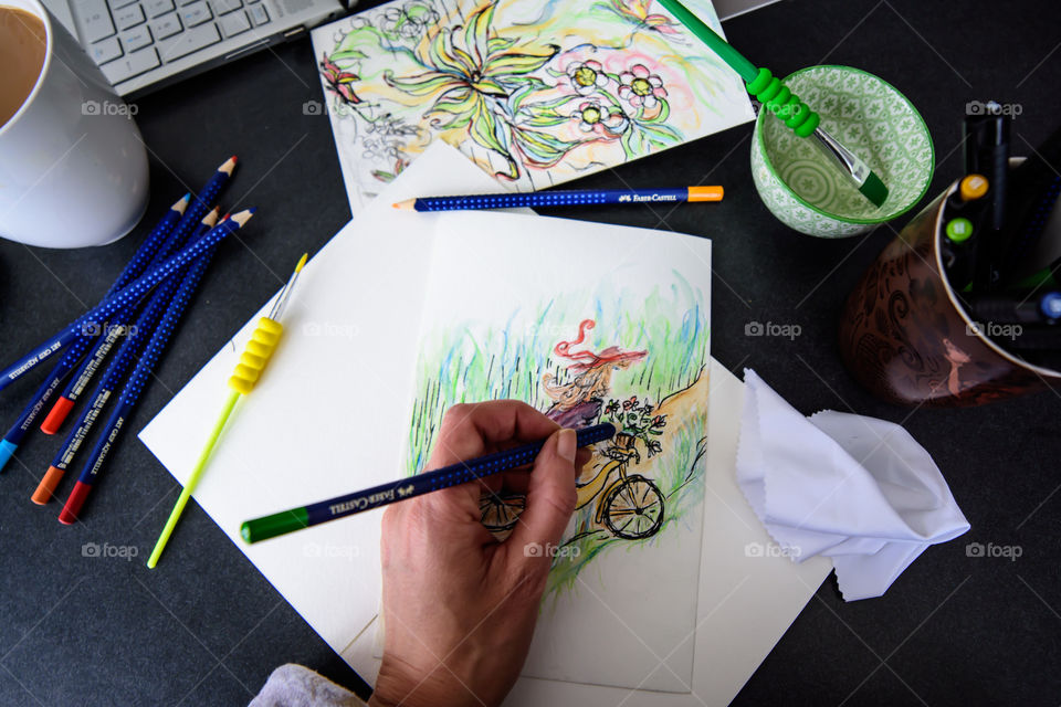 Flat lay Closeup of hand holding Faber-Castell Aquarelle Colored watercolor pencils sketching girl on bicycle in countryside with sketchbook on table with laptop and coffee conceptual work life balance, creativity, hobby artist lifestyle photography