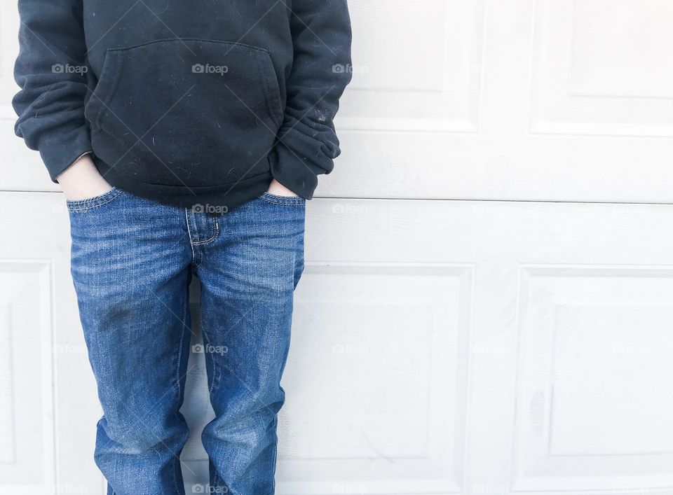 Boy with hands in the pockets of his blue jeans wearing a black hooded sweater standing next to a wall 