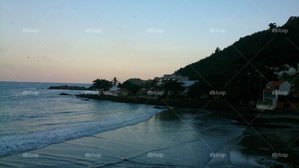 Evening view of the ocean.  Houses on the mountainside, rocks, horizon, waves and surf, blue skies.