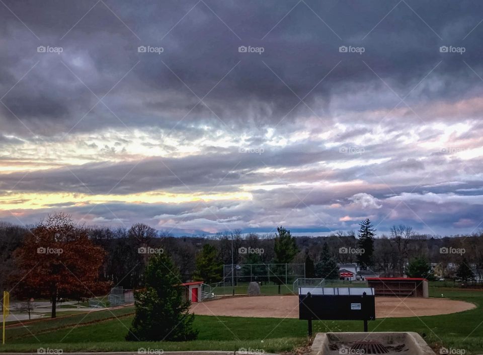 Sun setting behind pink, purple and gray cloud formations in a small Midwest town in Michigan over an empty baseball field at a high school which is surrounded by forest trees