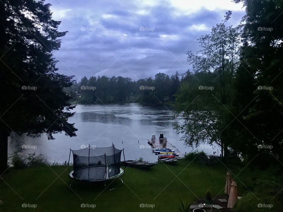Beautiful view of a lake from a lake house with a dock and a large backyard with a trampoline and cloudy skies