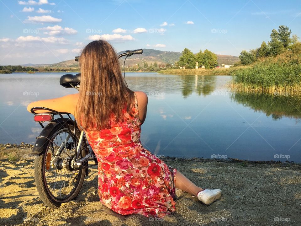 Back of woman wearing flower dress sitting on the sand looking at the lake with bicycle near her