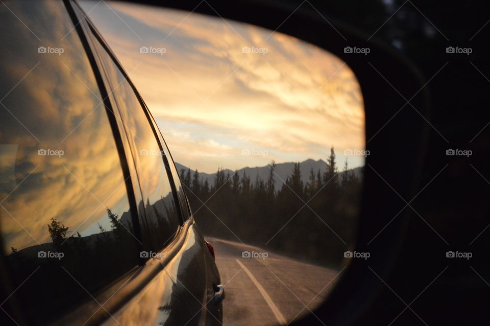 Colorful sunset in the side rear mirror.  Lovely view which includes pine trees and mountains.  Drive through the Canadian Rockies. 
