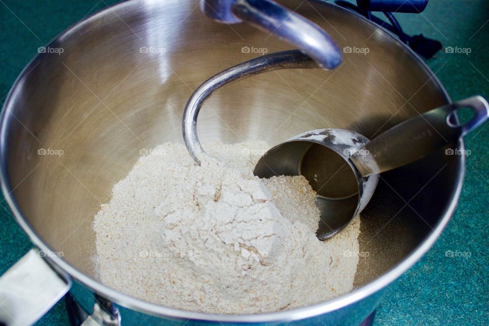 Whole-grain four and stainless steel measuring cup in mixing bowl of standing mixer fitted with dough hook on green countertop 