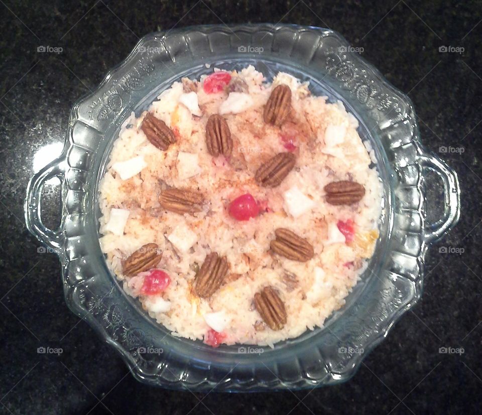 homemade ambrosia marshmallow salad in a clear blue depression glass serving bowl with handles.  it is garnished with maraschino cherries, pecans and shredded coconut. it sits on a dark granite countertop surface .