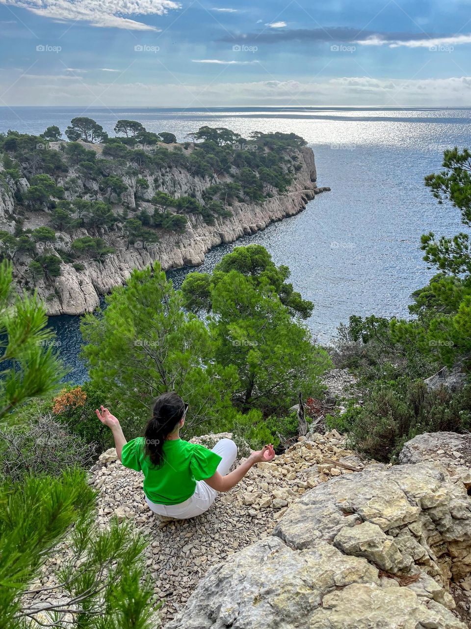 A young caucasian brunette girl in a green t-shirt and white jeans sits from the back meditating on the ridge of a high mountain with a panoramic landscape view of the mediterranean sea in marseille, france, top view close-up.