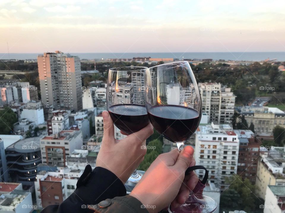 Two hands each holding a crystal glass coming together to make a toast with the buildings of the city of Buenos Aires in the background on a sky that embraces the sunset and floats on the river.