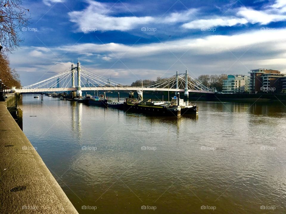 The murky Thames river in London is offset by a gorgeous blue, cloudy sky and a cool bridge off in the distance. A barge meanders down the waterway. 