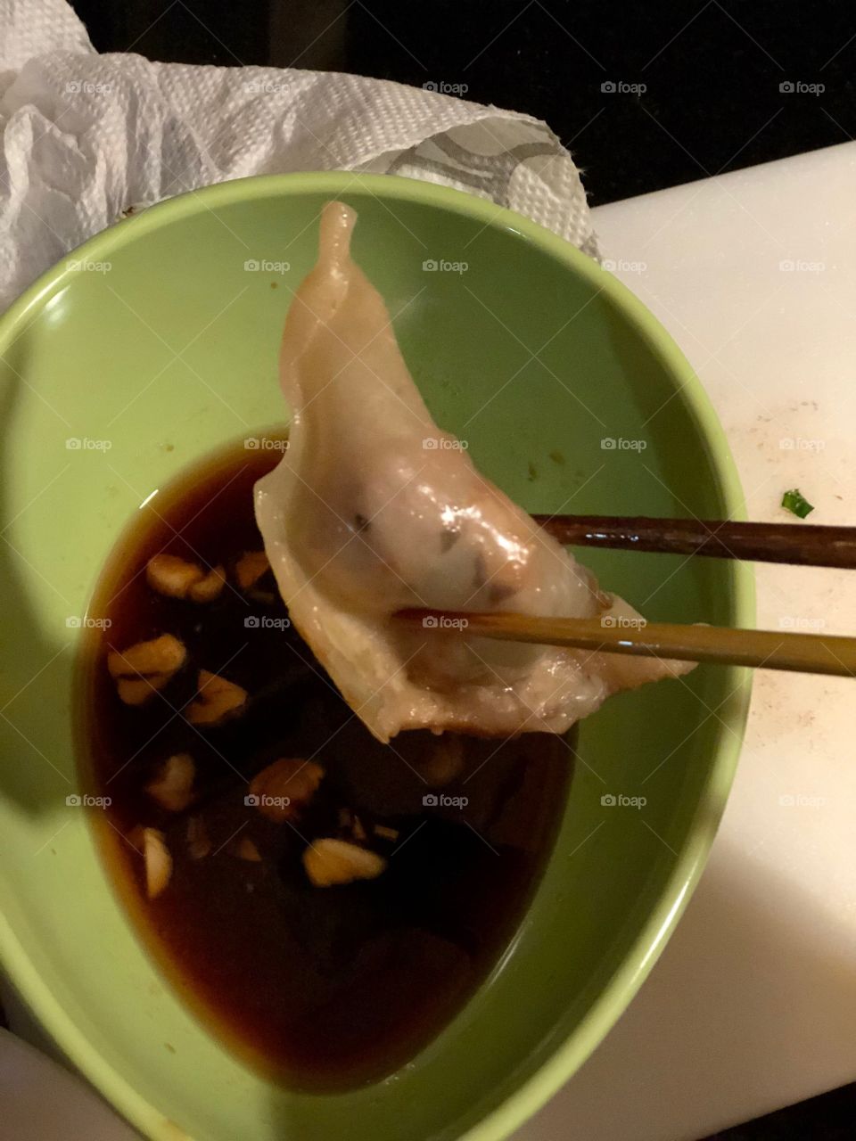 Chopsticks holding a potsticker over a green bowl filled with soy sauce and garlic pieces. 