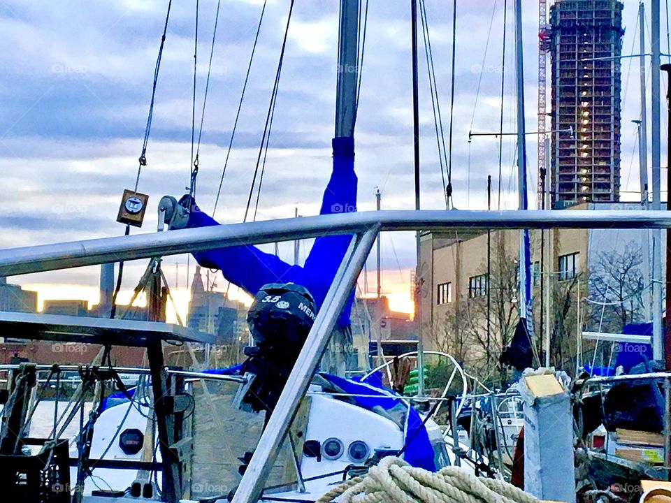 An assembly of objects: Our current boat “Salvation” 2’s railing, rope & motor gear, masts, ropes, mast & blue sail cover of the boat in front of us, the wall by the shore, building w scaffolding, houses afar. 2021. Hypnotic Productions