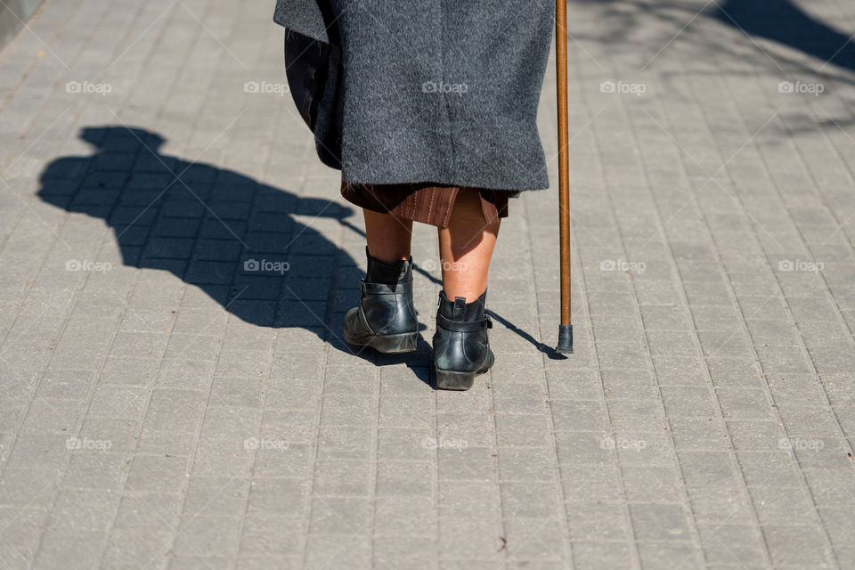 On a sunny day old woman walking down the street with walking stick. Womans shadow is visible on the sidewalk. View from back.