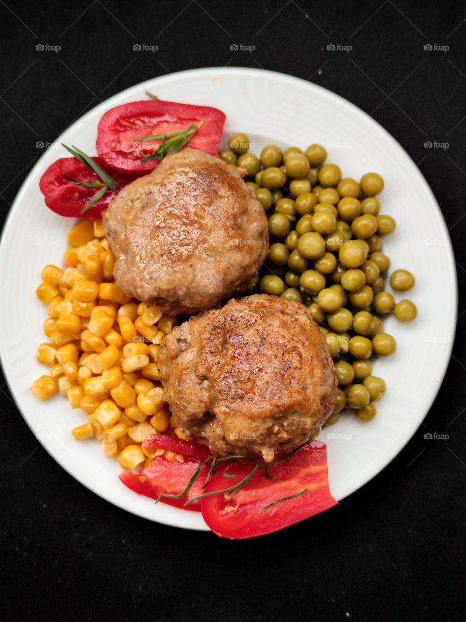 White plate on which are laid out two meat patties, corn grains, green peas, tomato slices with chopped herbs
