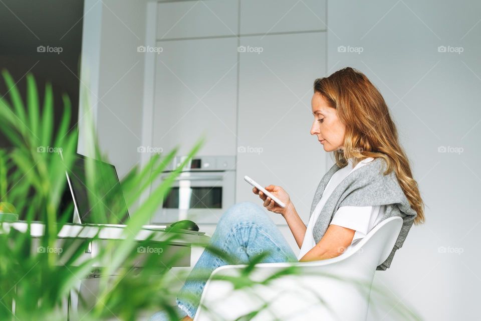 Young adult forty years beautiful blonde woman with long hair working on laptop using smartphone on kitchen at home, remote work, video call, freelancer
