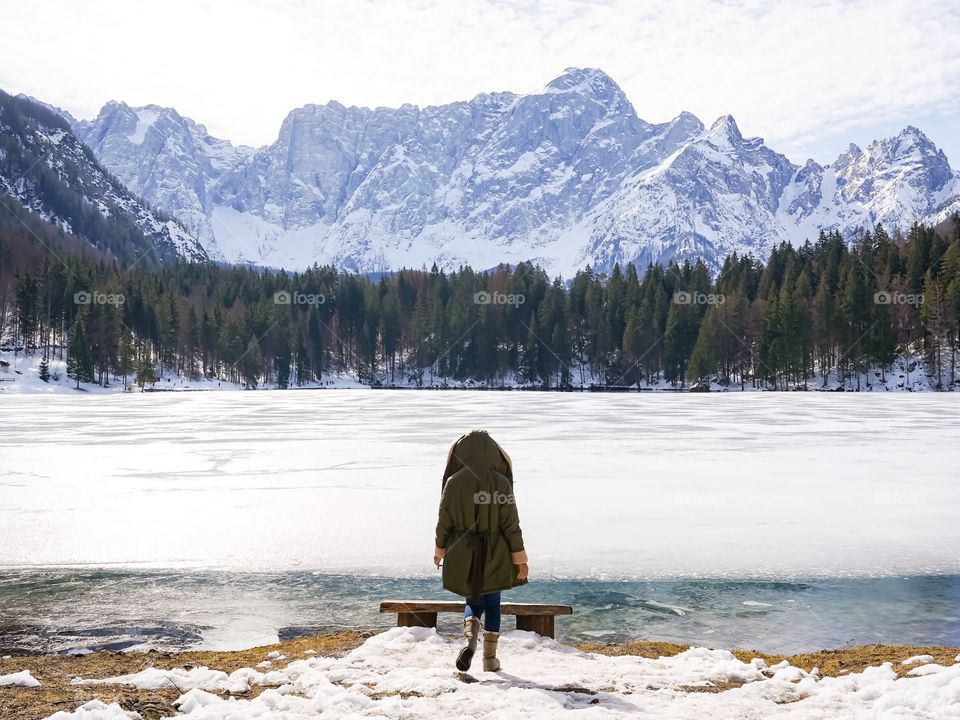 People from behind walking to the bench by icy lakeside in wintertime with incredible view of snowy mountains at front