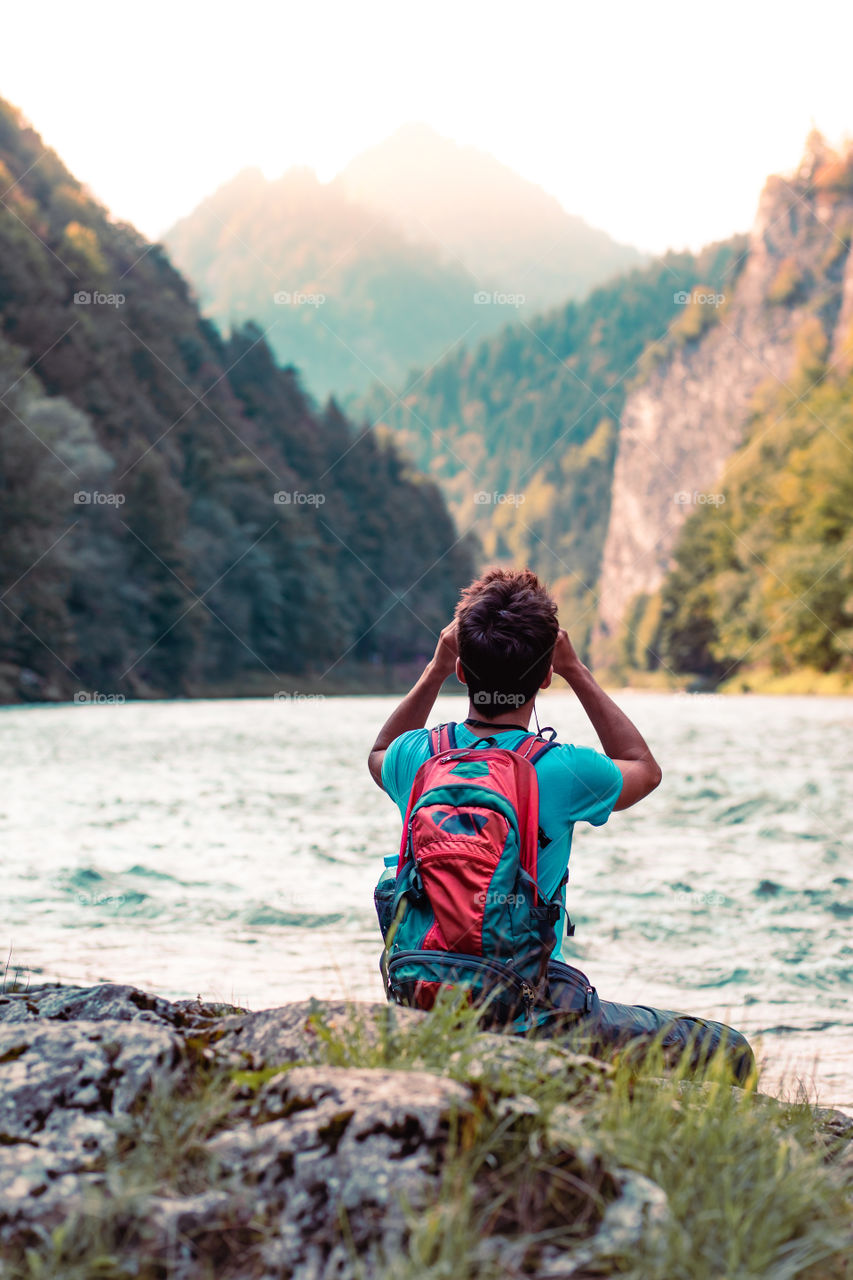 Young tourist with backpack looks through a binoculars on mountains peaks, stands on a rock over a river. Boy spends a vacation in mountains, wandering with backpack, he is wearing sports summer clothes