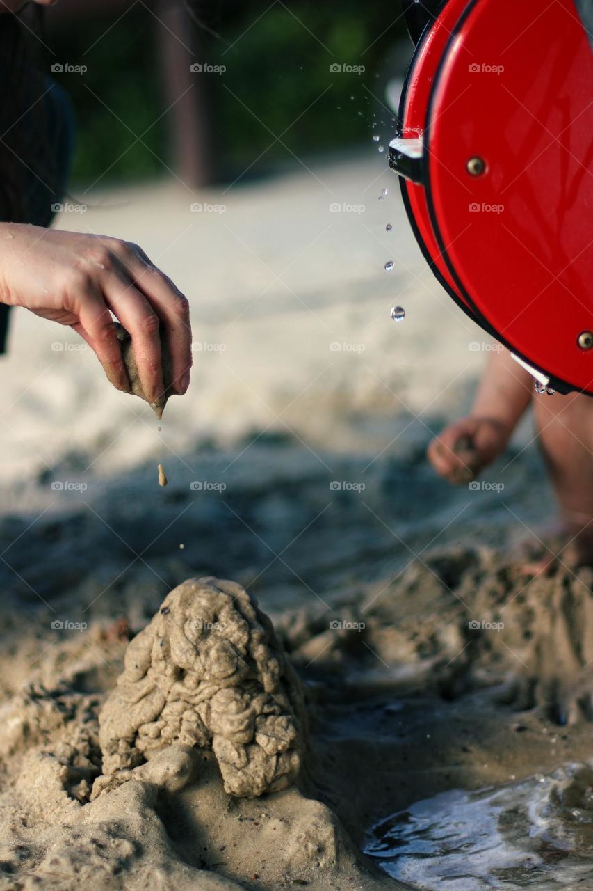 Hand of a woman and a toddler building a sandcastle with sand and water while drops of water drip from the water wheel