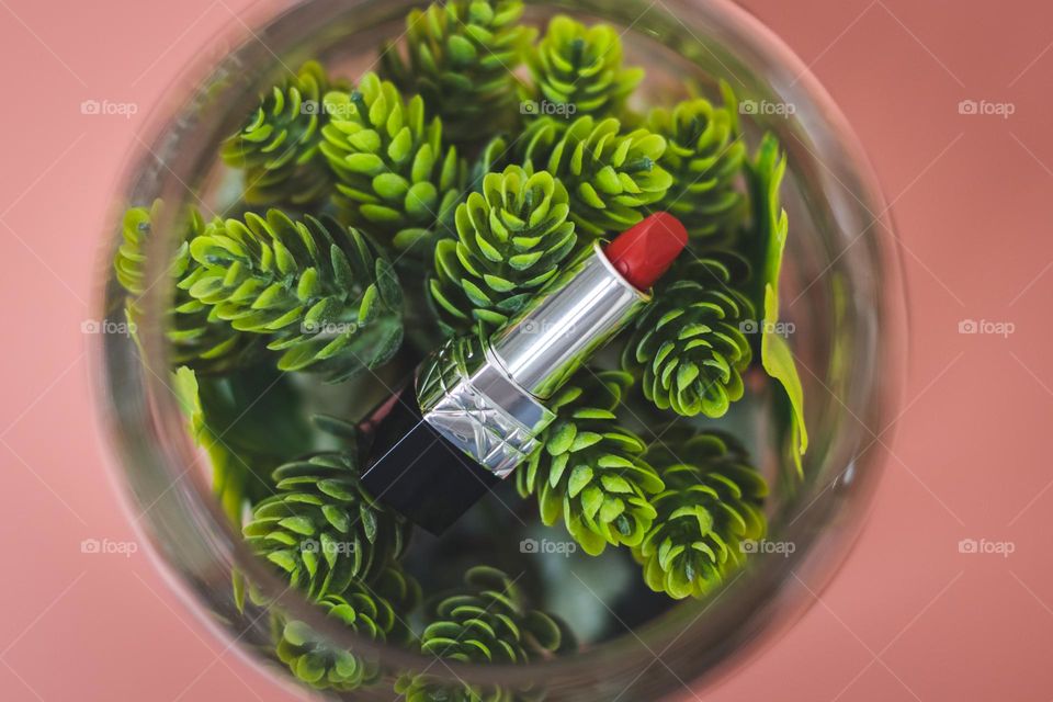 One red lipstick lies in a glass jar on a green flower on a pink background, flat lay. Concept cosmetics female, beauty salon.