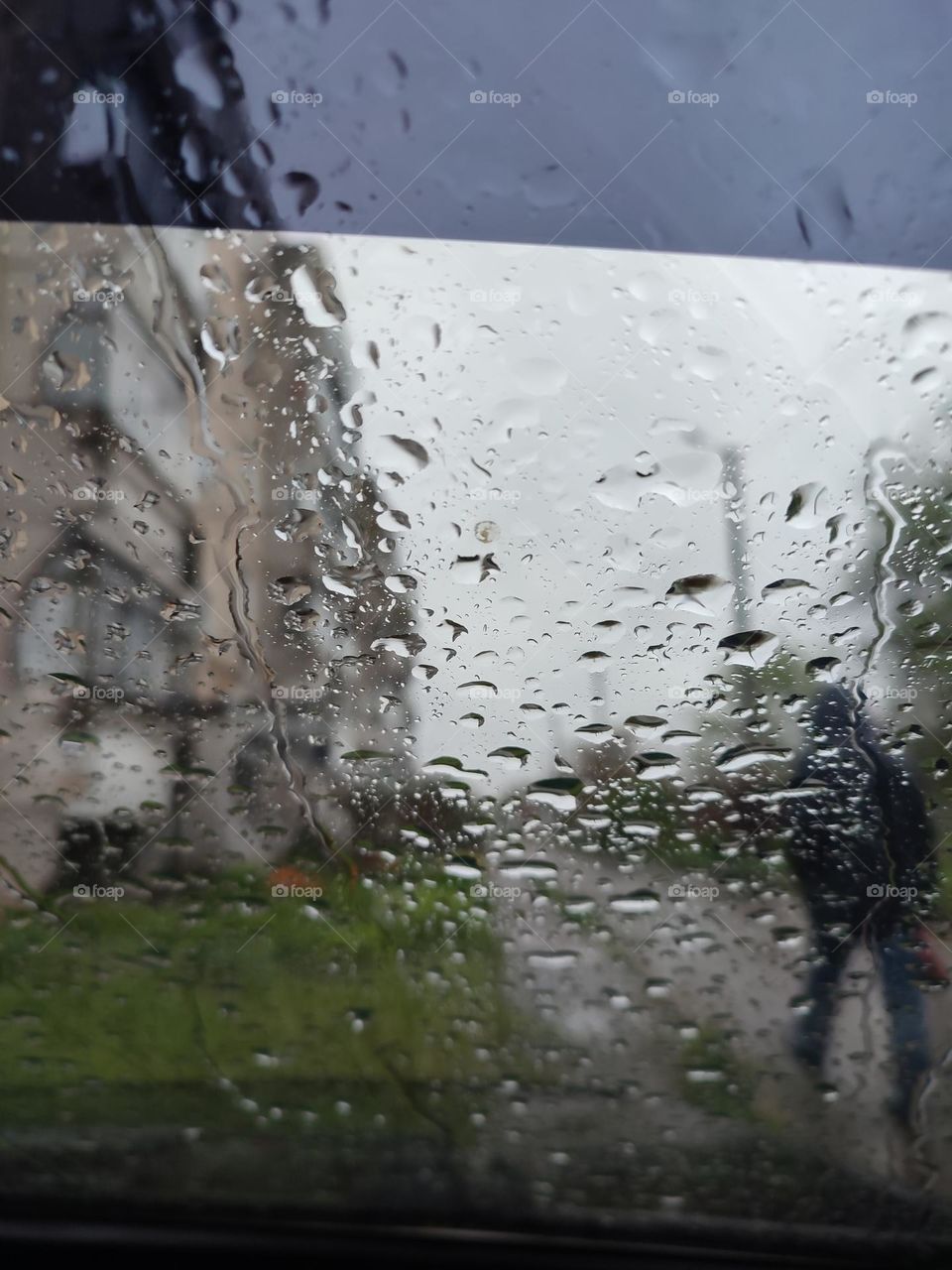 Raining day in the town, view from the car. Man walks in the rain. Raindrops running on the car window glass. Blurred skyline, multi storey building, courtyard, wet road. Spring thunderstorm. View from the window, wet weather. Walking person