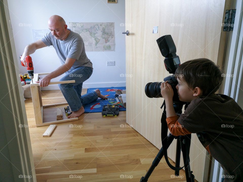 Boy behind camera taking picture of his parent assembling furniture.