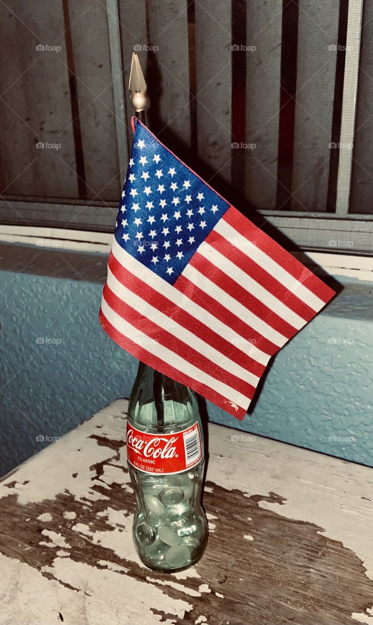 A small American flag rising out of a small glass Coca-Cola bottle resting on a weathered white wooden table in front of a blue wall and a window